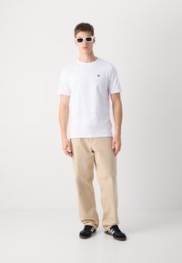 Hvid bomulds t-shirt med et lille sort logo, beige løstsiddende bukser og sorte sneakers med hvide striber. Model iført hvide solbriller.