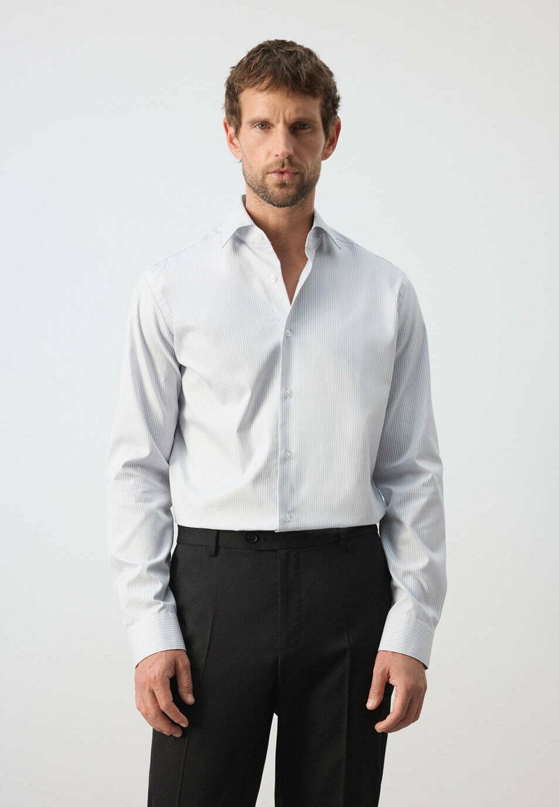 Homme portant une chemise à rayures claires boutonnée, rentrée dans un pantalon de ville noir, se tenant devant un fond clair uni.