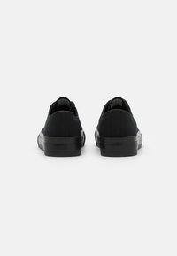 Black canvas sneakers with a low profile, rubber sole, and textured fabric. Green branding on the tongue, simple design, no visible patterns.