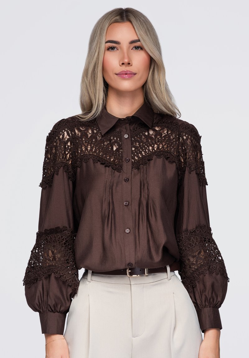 Femme aux longs cheveux blonds portant un chemisier marron foncé avec des détails en dentelle et un pantalon beige clair taille haute sur un fond uni.