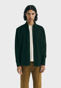 Tmavě zelená button-up košile vyrobená z měkkého materiálu, s kapsou na hrudi a volným střihem, doplněná o světle hnědé kalhoty.