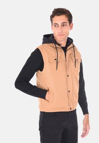 Giubbotto gilet con zip di colore tan, con texture a coste, caratterizzato da tasche sul petto doppie, cappuccio nero e maniche lunghe nere a contrasto. Le tasche sono dotate di bottoni a pressione.