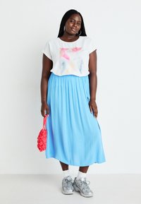 White short-sleeve top with a colorful graphic design, paired with a blue pleated maxi skirt and silver sneakers. Holding a red textured handbag.