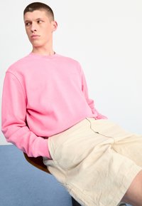 Rosa Sweatshirt mit langen Ärmeln und Rundhalsausschnitt, kombiniert mit hellbeigen Shorts. Das Sweatshirt hat eine lockere Passform und dezente Markenkennzeichnung.