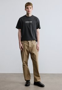 Tumši pelēka kokvilnas T-krekls ar baltu tekstu, kopā ar gaiši brūnām taisnām biksēm un melniem apaviem. Minimālistisks dizains ar vienkāršām krāsām.