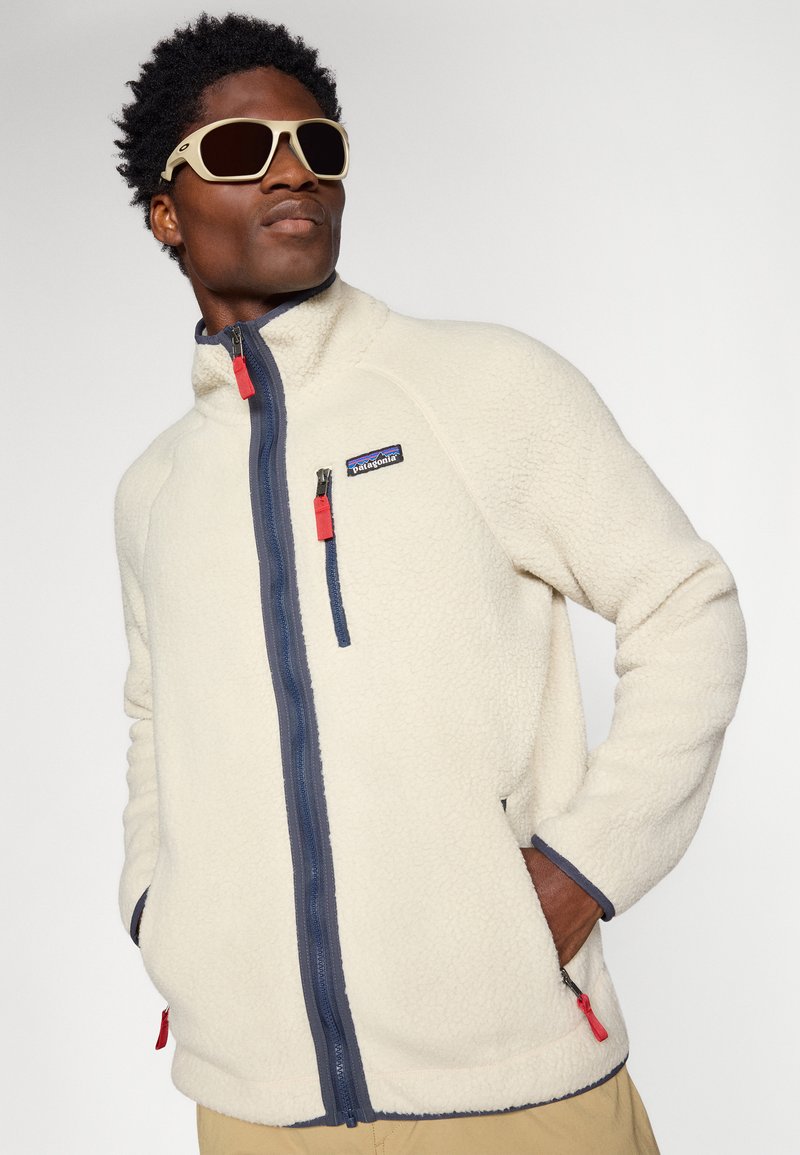 Giacca in pile color crema con cerniera blu navy e dettagli rossi, dotata di un colletto alto, tasca sul petto con zip e tasche laterali.