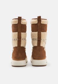 Botas de ante marrón con un borde de piel sintética crema, que cuentan con una parte superior acolchada, cordones ajustables y una suela de goma blanca con el logo "GUESS" grabado.