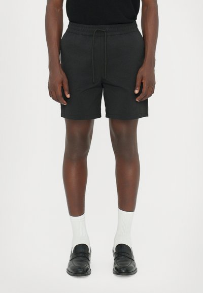 Pantalones cortos negros con una cintura elástica y cuerda de ajuste. Llevados con calcetines tobilleros blancos y mocasines negros. La tela parece suave y ligera.