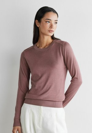 Kobieta o długich ciemnych włosach, ubrana w wrzosowy sweter z długim rękawem i białe spodnie, stojąca na tle jednolitej białej ściany, patrząca w prawo.