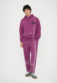 Hanorac mov cu buzunar frontal și logo, asociat cu pantaloni mov asortați. Design cu glugă și un gradient subtil, cu detalii de șnur. Pantofi casual.
