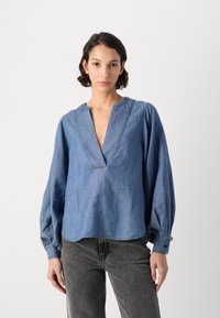 Blå denimbluse med dyp V-hals, rynkede lange ermer og subtile stikninger. Kombinert med mørkegrå bukser.