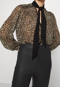 Blusa de leopardo transparente en beige y marrón, con una corbata negra al cuello, hombros fruncidos y mangas largas, combinada con jeans negros ajustados.