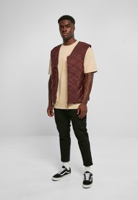 Körsbärsröd kviltad väst med dragkedja, bärs över en beige t-shirt, kombineras med svarta byxor och svarta sneakers med vita accenter.