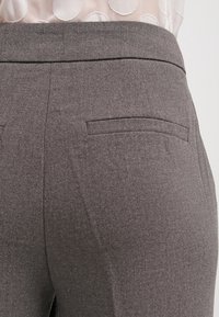 Pantalon gris sur mesure en tissu lisse et texturé, avec une taille ajustée et deux poches arrière. Des coutures discrètes soulignent le design.