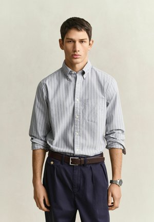 Jeune homme portant une chemise à rayures bleu clair avec boutons et manches retroussées, pantalon bleu marine, ceinture marron et montre-bracelet argentée, debout devant un fond uni.