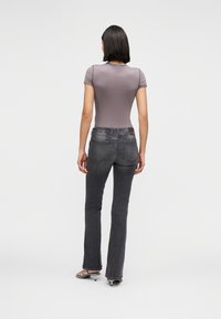 Γυναίκα με κοντά σκουρόχρωμα μαλλιά, που φοράει εφαρμοστό γκρι μπλουζάκι, ξεθωριασμένο μαύρο τζιν και ασημένια ψηλοτάκουνα σανδάλια, στέκεται γυρισμένη με την πλάτη σε λευκό φόντο.