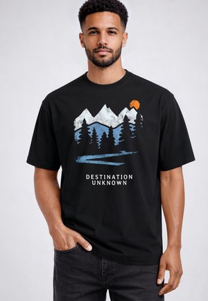 Mann trägt schwarzes T-Shirt mit Berg-, Baum-, Flussgrafik und orangefarbener Sonne, Text „DESTINATION UNKNOWN“, steht mit der Hand in der Tasche.