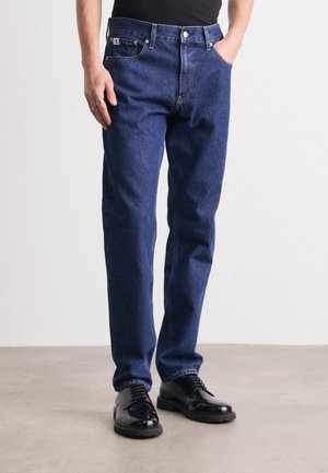 Mann trägt dunkelblaue Jeans, schwarze Lederschuhe, schwarzes Oberteil, steht mit einer Hand in der Tasche auf hellgrauem Boden.