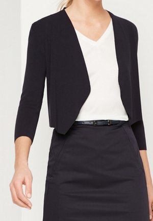 Femme portant un blazer court noir sur un top blanc à col en V, associé à une jupe noire taille haute et une fine ceinture noire, partie médiane visible.