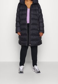 Schwarze Pufferjacke mit Kapuze, ausgestattet mit einem gesteppte Design, einem Frontreißverschluss und seitlichen Taschen, getragen über einem helllila Oberteil und schwarzen Hosen.