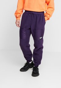 Purpurowe dresy z polaru z kontrastującymi purpurowymi nylonowymi kieszeniami, elastycznym pasem i ściągaczami. Czarne sneakersy dopełniają całość.