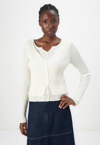 Ribbet hvit cardigan med dyp v-hals, knappestolpe og lange ermer. Kombinert med et denimskjørt med synlig sømdetalj.