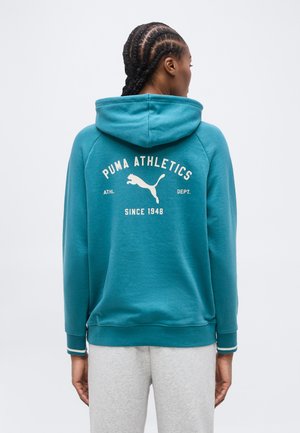 Persona con capelli intrecciati che indossa una felpa con cappuccio Puma color teal con la scritta "Puma Athletics Since 1948" e il logo, vista da dietro.