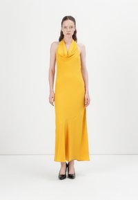 Robe jaune à col halter avec décolleté en cowl et ourlet asymétrique, en tissu lisse, associée à des talons hauts noirs.