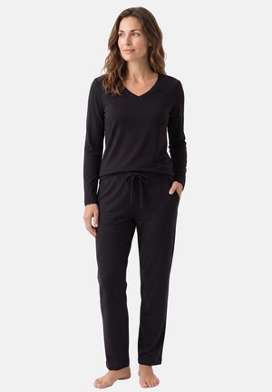 Donna in piedi a piedi nudi che indossa un top nero a maniche lunghe con scollo a V e pantaloni neri con coulisse, guarda leggermente di lato.