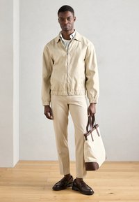 Μπεζ ελαφρύ μπουφάν με ελαστική μέση, σε συνδυασμό με κρεμ παντελόνι και καφέ loafers. Κρατάει μια κρεμ τσάντα tote με καφέ λεπτομέρειες.