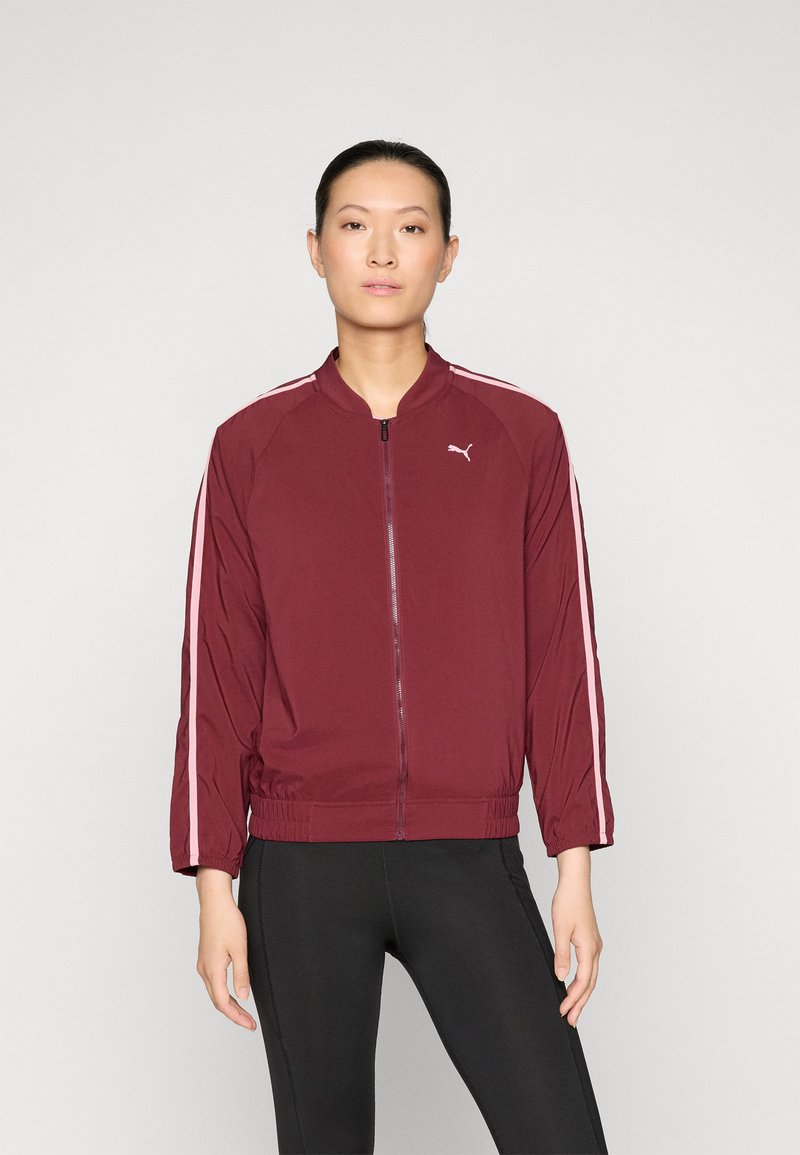 Bordeauxfarbene, leichte Bomberjacke mit rosa seitlichen Streifen, Reißverschluss vorne, elastischem Bund und elastischen Manschetten. Das Modell trägt dazu schwarze Leggings.