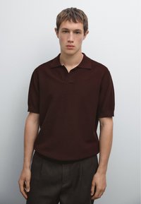 Polo a maniche corte realizzato in tessuto borgogna testurizzato con colletto e scollatura a V, indossato con pantaloni scuri su misura.
