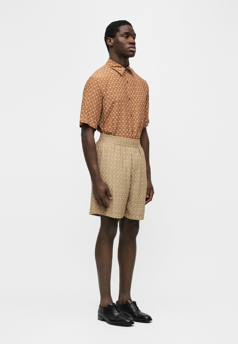 Mann steht in einem kurzärmeligen Hemd mit Muster und passenden beigen Shorts mit Muster, trägt schwarze Anzugschuhe, auf weißem Hintergrund.