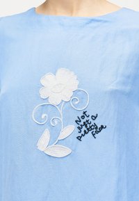 Lyseblåt stof med hvidt broderet blomstermønster og sort broderet tekst, der lyder "Ikke bare et smukt ansigt" nær halsudskæringen.