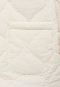 Materialul de quilt cu o nuanță deschisă de crem prezintă o mică buzunare cu margini rotunjite. Textura pare moale, cu modele de valuri rotunjite brodate pe întreaga suprafață.