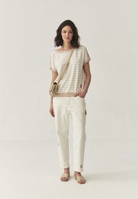 T-shirt rayé beige et crème à manches courtes, associé à un pantalon blanc avec des détails brodés. Le modèle porte un petit sac bandoulière beige.