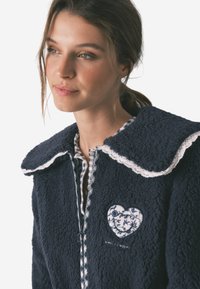 Giacca in pile blu navy con un grande colletto bianco bordato in pizzo e un'appliqué floreale a forma di cuore, con una fodera interna a quadri.