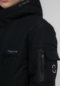 Svart vinterjacka med en dragkedjeförsedd ficka på ärmen, med en logotyp-patch och texturerad tyg. Justerbar huva och en stilren design.