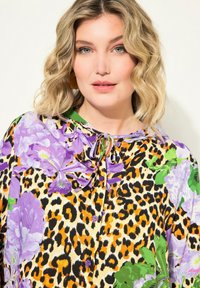 Květinová blůza v purpurové, zelené a černé leopardí potisku. Má zapínání na knoflíky vpředu, kravatu na krku a živé květinové vzory.