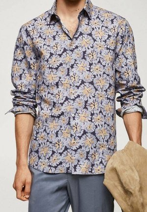 Homme portant une chemise fleurie à manches longues boutonnée avec les manches retroussées, tenant une veste beige dans une main.