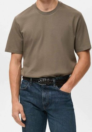 Man draagt een olijfgroen kortärmelig overhemd, ingestopt in een blauwe spijkerbroek met een zwarte gevlochten riem, met één hand in de zak tegen een witte achtergrond.