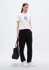 Λευκό βαμβακερό μπλουζάκι με μοβ λογότυπο, συνδυασμένο με μαύρη φόρμα με κορδόνι στη μέση και λευκά αθλητικά παπούτσια. Μαύρη τσάντα tote κρατημένη στο χέρι.