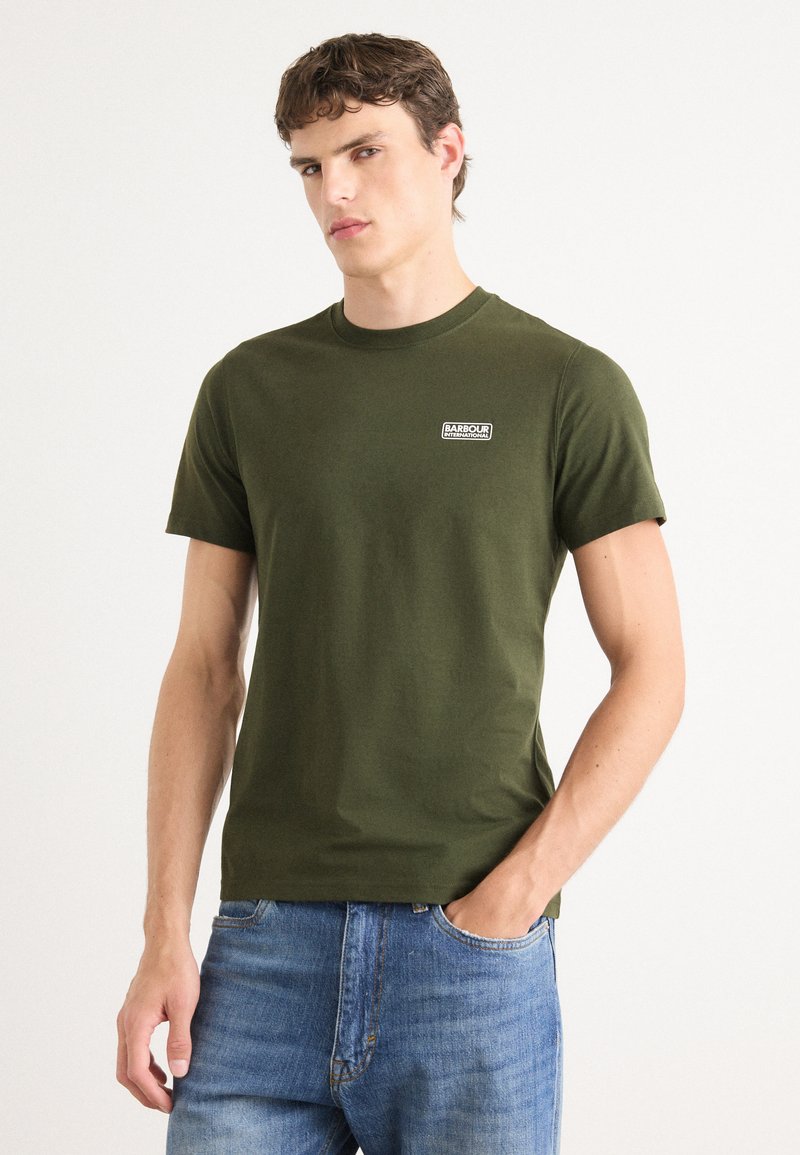 Σκούρο πράσινο βαμβακερό t-shirt με στρογγυλή λαιμόκοψη και κοντά μανίκια, διαθέτει ένα μικρό άσπρο ορθογώνιο λογότυπο στο αριστερό στήθος. Φοριέται με μπλε τζιν.