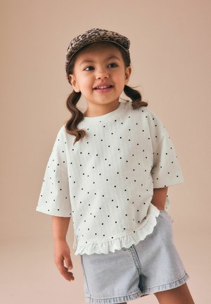 Junges Mädchen mit Zöpfen, das eine Leopardenmütze, ein weißes gepunktetes Hemd mit Volantsaum und helle Jeansshorts trägt, lächelnd und posierend.