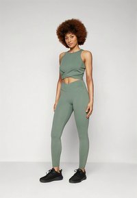 Frau mit lockigem Haar, die ein grünes ärmelloses Crop-Top und hochgeschnittene Leggings trägt, steht mit schwarzen Turnschuhen vor einem einfarbigen Hintergrund.