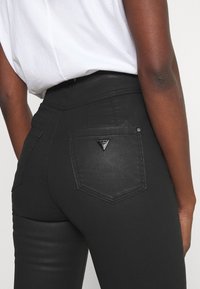 Primer plano de una persona que lleva pantalones ajustados negros con un pequeño logo metálico triangular en el bolsillo trasero derecho y una camisa blanca metida.