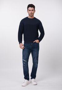 Homme portant un pull bleu marine, un jean bleu déchiré et des baskets blanches, debout avec une main dans la poche, devant un fond uni.