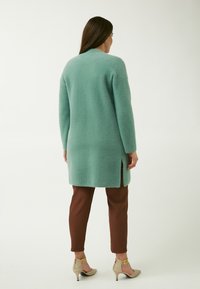 Lang, mintgrøn, fuzzy cardigan med høj hals og sideslidser, parret med brune bukser og beige højhælede sko.