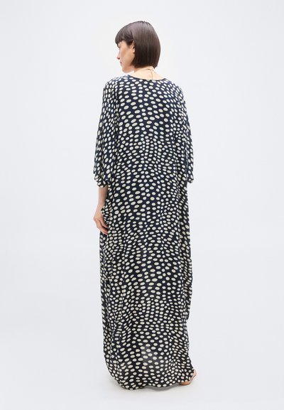 Γυναίκα που στέκεται ξυπόλητη, γυρισμένη προς τα πίσω, φορώντας ένα μακρύ, φαρδύ μπλε φόρεμα με ακανόνιστα άσπρα πουά και μανίκια μέχρι τον αγκώνα.