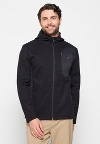 Zwarte fleece jack met ribbelstructuur, volledige ritssluiting, verstelbare capuchon en een ritszak op de borst met een logo aan de linkerzijde.